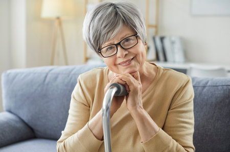 Portrait of happy grey haired woman posing with walking stick, focused at camera and smiling, posing on comfortable sofa, enjoying cozy atmosphere at home. Pension and retirement conceptの写真素材