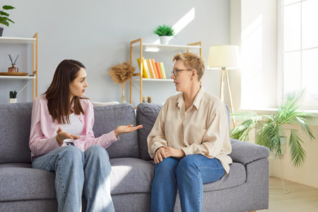 Young woman and senior woman sitting together on sofa at home, having serious conversation. Elderly mother listening to adult daughter expressing concern, sharing thoughts and actively gesticulating.の写真素材