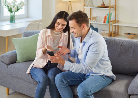 Cheerful young couple using smartphone sitting on sofa in living room at home browsing apps. Young man showing video or news on mobile phone to his smiling wife. People and technology concept.の写真素材