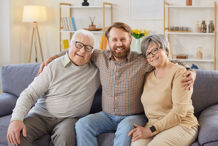 Portrait of happy elderly parents with adult son embracing together at home, showcasing family love in a cozy setting filled with joy and connection in a warm domestic environment.の写真素材