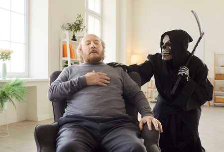 Portrait of senior old sick man sitting on chair suffering from heart attack and chest pain being approached by death in grim reaper costume holding scythe. Retired male person at home.の写真素材