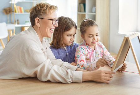 Portrait of happy smiling senior grandmother sitting at the desk with her little grandchildren playing in educational game. Kids with granny spending time together at home. Leisure and people conceptの写真素材