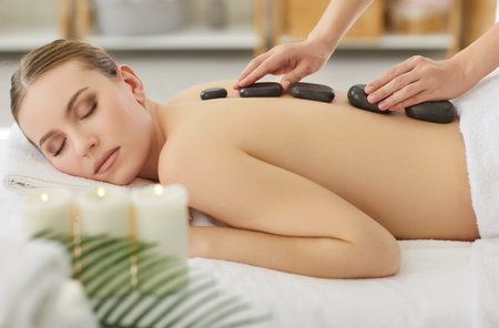 Calm young woman lying on massage table with eyes closed, receiving relaxing hot stone therapy from masseur in spa center, surrounded by lit candles creating calm and peaceful wellness atmosphere.の写真素材