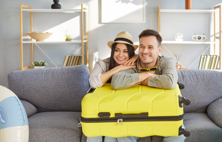 Happy young couple sitting together on sofa with packed suitcase on their knees, ready for summer journey. Smiling husband and wife hugging, feeling excited for upcoming trip. Traveling and vacation.の写真素材