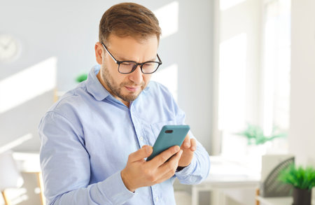Portrait of focused man in glasses standing in bright room or office, closely examining smartphone screen with serious expression. Adult male analyzing or reading something important on his device.の写真素材