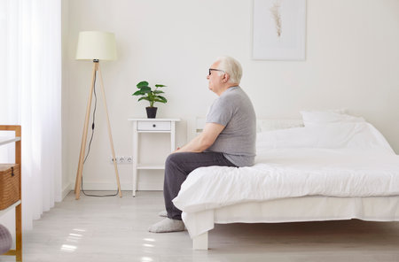 Grandfather unhappy alone at home, senior old man sitting on bed, sleepless mature patient suffering from insomnia, health problems, feeling lonely, missing resident of nursing homes in hospitalの写真素材