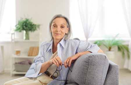 Smiling concentrated attractive senior portrait, aged female holding glasses, pleasant confident old age woman at home, pensive mature lady , thinking, planning in living room alone, sitting on sofaの写真素材