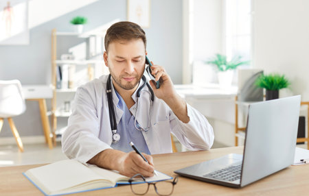 Concentrated male doctor or nurse handling a phone call while working with a laptop in a hospital. Online communication and delivering healthcare expertise through digital tools or internet.の写真素材
