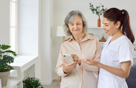 Young friendly smiling female nurse showing elderly woman how to use smartphone. Caregiver teaching senior person to handle with mobile phone. Nursing home service and patient support concept.の写真素材