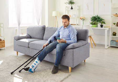 Young disabled man in casual clothes sitting on sofa at home with crutches holding mobile phone in hands. Guy with injured leg in plaster or splint with walking stick using smartphone.の写真素材