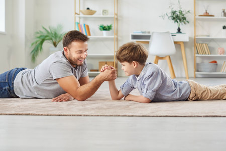 Portrait of happy cheerful father arm wrestling with little boy son lying on floor in living room at home. Young man parent having fun playing with child indoors enjoying time together.の写真素材