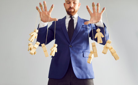 Bottom view of a young businessman puppeteer wearing suit controlling wooden human figures marionettes by hands hanging on rope isolated on white background. Manipulating and people control concept.の写真素材