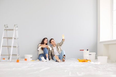 Smiling happy couple painting wall of new home sitting near ladder. Married man and woman sitting on floor and planning repair renovation preparing to move into new flat. Moving day conceptの写真素材
