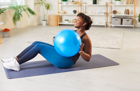 Sporty fit middle aged happy african american woman doing fitness practice at home on mat, beautiful female working out doing exercise for abs with blue Swiss ball during sport class, trainingの写真素材