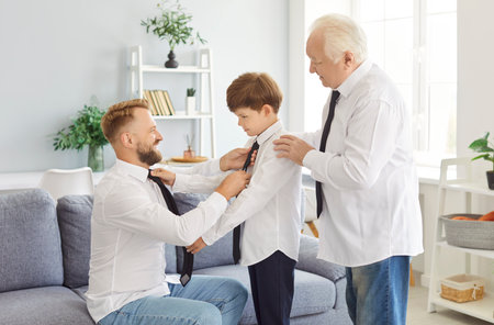 Cheerful multi generation male family wearing white shirts dressing, fixing outfits and preparing for special occasion. Smiling father and grandfather helping young boy, teaching him to tie necktie.の写真素材