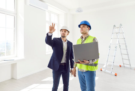 Portrait of young foreman discussing with professional architect design project or repair of new apartment on a laptop standing in empty room. Meeting with builder about interior decoration.の写真素材
