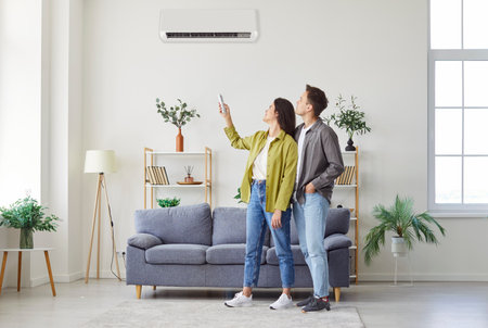 Young couple turning on air conditioning system in living room. Happy man and woman using remote control to change temperature on their white AC on the wall, while standing at home. Climate control.の写真素材