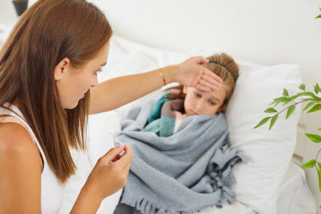 Portrait of worried young mother checking temperature with thermometer of her sick little child feeling bad with influenza lying on bed at home suffering from flu or cold. Healthcare concept.の写真素材
