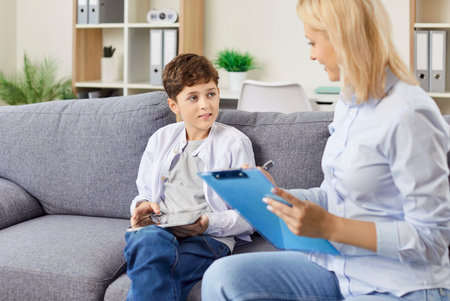 Portrait of a friendly young female psychologist woman doctor talking with her child boy patient sitting on sofa in office during therapy session. Psychotherapy and kids mental health concept.の写真素材
