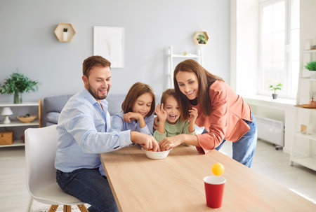 Young happy parents mother, father and two children having fun together in living room at home playing funny game with kids throwing balls into cardboard cup. Family and leisure concept.の写真素材