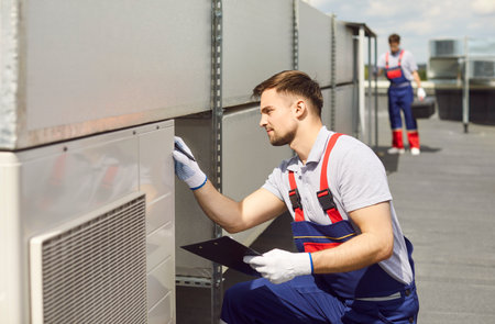 Worker doing inspection, testing and repairing of the air conditioning or ventilation system on the roof. Maintenance activity ensures the HVAC system operates, providing comfort indoors.の写真素材