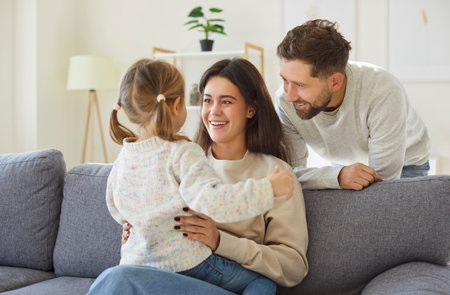 Happy family at home, young mother and father with cute little daughters sitting on couch, looking at each other, enjoying tender moment, smiling parents and child having fun together, playingの写真素材