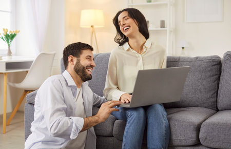 Happy laughing young couple watching laptop screen, funny movie, planning budget, reading good news in email, smiling woman and man looking at computer, checking finances, sitting at home togetherの写真素材
