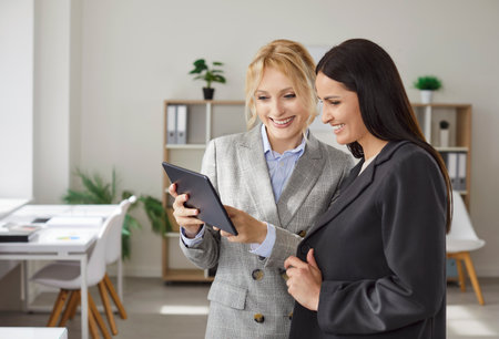 Two businesswomen collaborating together, focused in tablet, working on project, dressed formally, standing in cabinet, discussing something. Women colleagues using modern technologiesの写真素材
