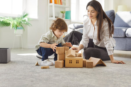 Mother with boy child enjoying playtime building tower with wooden cubes during leisure at home, fostering family togetherness in a cozy setting with focus on creativity and bonding.の写真素材