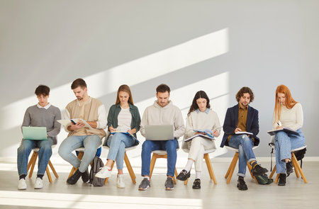 Focused young students sitting in row on chairs in room, reading books and working on laptops. Group of male and female job seekers waiting for job interview. Human resources, employment, education.の写真素材