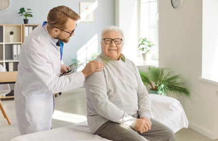 Senior happy man patient in medical clinic talking with male young friendly doctor wearing stethoscope sitting on couch in office during examination. Doctor support and health care concept.の写真素材