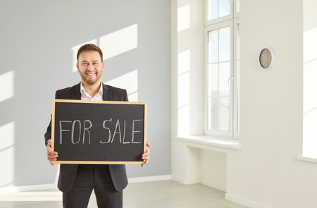 Portrait of happy smiling male real estate agent wearing formal suit showing a sign with inscription for sale standing in modern empty apartment selling house and looking at camera.の写真素材