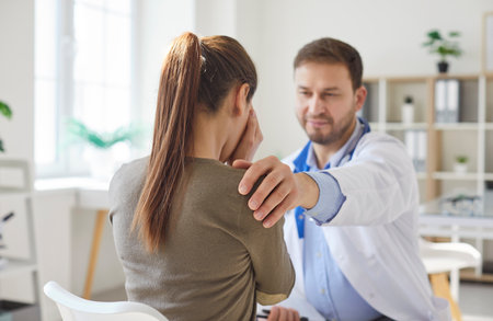 Male doctor support young upset unhappy sick female patient putting hand on her shoulder. Physician sitting in medical clinic giving consultation a woman during medical examination in office.の写真素材