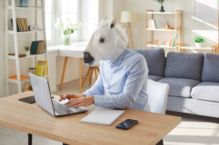 Funny extravagant happy man wearing horse mask working at laptop online successful job at workplace table, looking at display, typing, thinking on online content, using computer workの写真素材