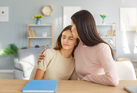 Young cheerful happy mother hugging and kissing her teen school girl daughter sitting at desk at home tired from school homework. Woman support child in studying. Education concept.の写真素材
