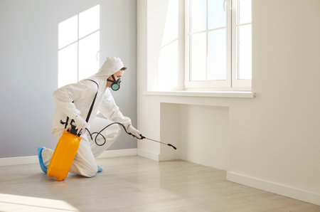 Professional exterminator in protective white suit and gas mask spraying insecticide on floor in empty room. Male pest control worker with sprayer bottle using chemical treatment for disinfection.の写真素材