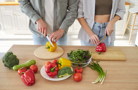 Man and woman engage in food preparation, cutting colorful vegetables on wooden boards. Angled top view of hands of people cooking together at home, demonstrating teamwork in creating healthy meal.の写真素材