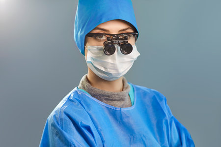 Portrait of professional confident female doctor surgeon wearing blue medical uniform and binocular loupes looking at camera at hospital. Medicine, surgery and emergency help concept.の写真素材