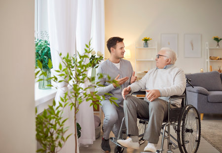 Young friendly male social worker talking with senior elderly disabled man with disability sitting in wheelchair. Cheerful nurse or caregiver with handicapped patient in nursing home.の写真素材