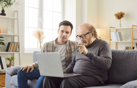 Young man, adult son teaching interested mature senior father using laptop computer online applications at home, watching photo video content in social network, shopping in internet store, modern techの写真素材