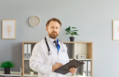 Experienced man doctor standing confident in modern hospital office, writing down notes as holding clipboard, taking care of his patients health, giving medical help. People and health conceptの写真素材