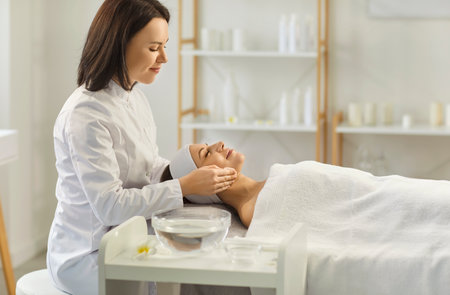 Cheerful female beautician making facial treatment on woman client face in spa salon. Relaxed pretty young woman receiving beauty cosmetology procedure. Skin care and wellness concept.の写真素材