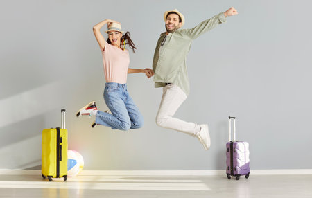 Funny excited happy couple in hats having fun ready for holiday trip. Joyful smiling man and woman tourists jumping with packed luggage on gray wall background. Vacation and summer journey concept.の写真素材