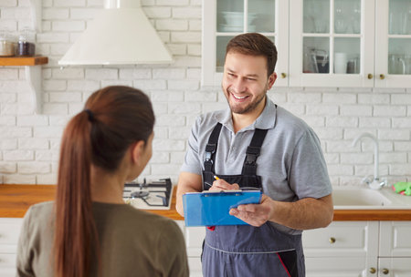 Man plumber talking with female client signing contract for plumbing services at home. Male mechanic or repairman consulting woman customer standing in kitchen. Maintenance and repair service.の写真素材