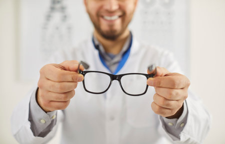 Man professional ophtalmologist holding spectacles with corrective lenses, taking care of your eye health, wearing white coat, ready to provide eye care to patients, giving prescriptionsの写真素材