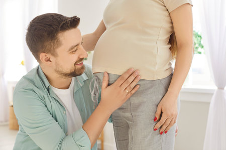 Portrait of young cheerful man looking at his pregnant womans tummy and smiling sanding in living room at home. Loving husband enjoying pregnancy of his wife. Couple expecting a baby.の写真素材