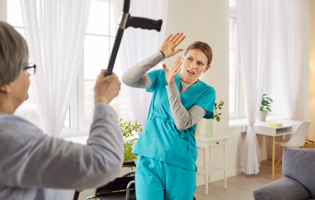 Old patient demonstrates her aggression towards nurse or caregiver, threatens with crutch, expresses rage, dissatisfied with care. Frightened female carer protects herself from angry womanの写真素材