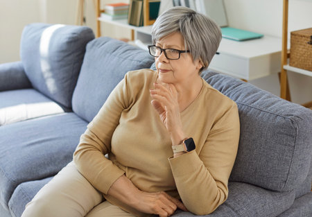 Elderly thoughtful woman wearing spectacles and comfortable clothes, sitting on sofa, contemplating about life, feeling cozy at home in living room. Old people and lifestyle conceptの写真素材