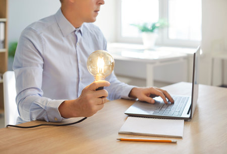 Cropped shot of male company employee sitting at desk, using laptop and holding glowing lightbulb in hand. Creative professional generating bright idea and working on innovative project in office.の写真素材