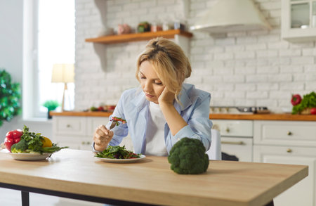 Dissatisfied woman eating vegetable salad while following diet at home in kitchen, focusing on healthy food in a challenging setting with dedication to wellness and nutrition every day.の写真素材